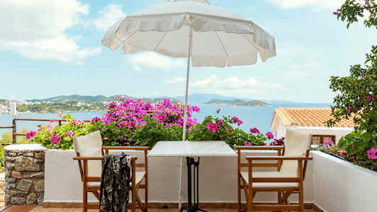 En uteplass med bord, stoler, en parasoll og blomster, med havutsikt. (AI-generert tekst)