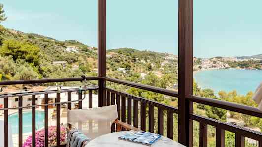 Utsikt fra en balkong med bord og stol mot et basseng, vegetasjon og fjord. (AI-generert tekst)