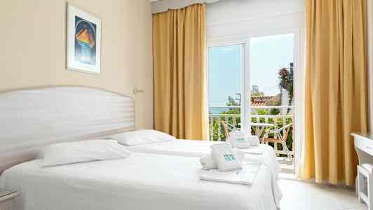 Et lyst hotellrom med to enkeltsenger, store vinduer med gulldraperier og utgang til en balkong. (AI-generert tekst)