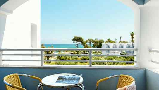 Utsikt fra en balkong med havet i bakgrunnen. En gul stol og et bord er på balkongen. (AI-generert tekst)