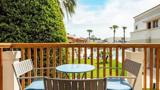 Utsikt fra en balkong med bord og stoler mot en hage og basseng. (AI-generert tekst)