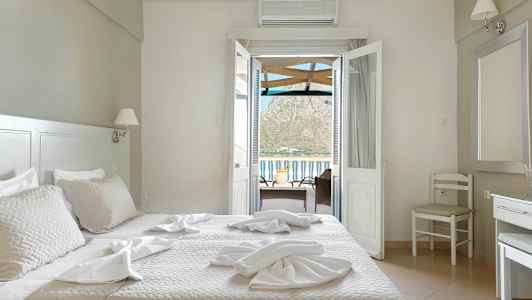 Et hotellrom med en seng, stoler og et bord med utsikt gjennom dørene mot balkongen og fjellene i bakgrunnen. (AI-generert tekst)