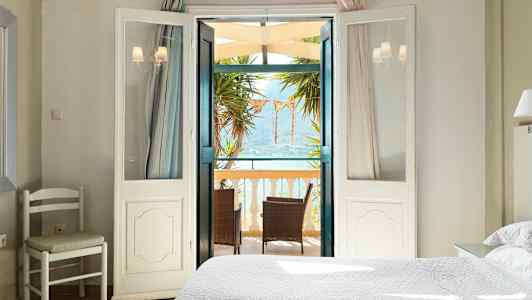 Et lyst soverom med åpne dører til en balkong med møbler og utsikt over havet. (AI-generert tekst)