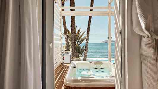 Utsikt fra et rom mot en terrasse med boblebad, palmer og hav. (AI-generert tekst)