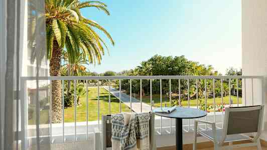 Utsikten fra en balkong med et bord, stoler og et håndkle, mot en hage med palmer og gangvei. (AI-generert tekst)