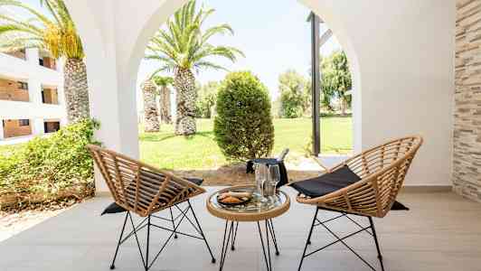 To flettede stoler og et bord med glass og en tallerken på en terrasse, med utsikt over en grønn hage med palmer. (AI-generert tekst)