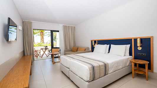 Hotellrom med en stor seng, sittegruppe og utsikt mot en terrasse og hage. (AI-generert tekst)