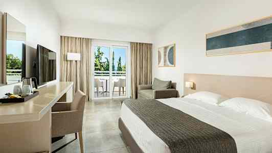 Et hotellrom med dobbeltseng, skrivebord og stol, lenestol samt en balkong med utsikt mot trær. (AI-generert tekst)