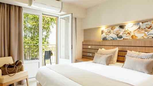 Et hotellrom med en stor seng, sittemøbel og åpen balkongdør med utsikt mot grønne trær. (AI-generert tekst)