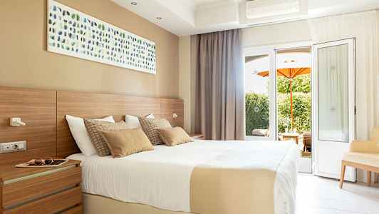 Et lyst hotellrom med en stor seng, veggkunst og altandører med utsikt over en hage med en oransje parasoll. (AI-generert tekst)