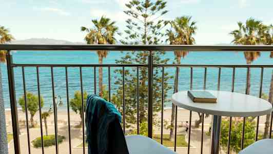 Utsikt fra en balkong med bord og stoler mot havet og palmer. (AI-generert tekst)
