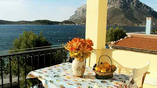 En utsikt fra en balkong mot havet, med et bord dekorert med en vase med oransje blomster og en kurv med sitroner. (AI-generert tekst)