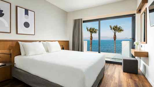 Hotellrom med hvit seng og utsikt over havet gjennom en glassdør. (AI-generert tekst)