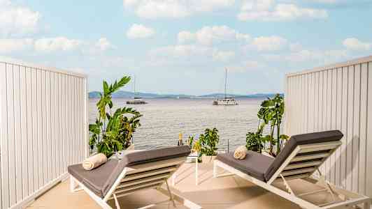 To solsenger med grå puter og håndklær plassert på en terrasse med utsikt over havet og seilbåter. (AI-generert tekst)