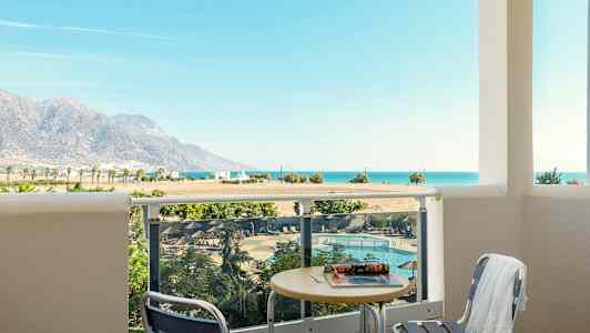 Utsikt fra en balkong med bord og stoler mot et basseng, landskap med fjell og havet i bakgrunnen. (AI-generert tekst)