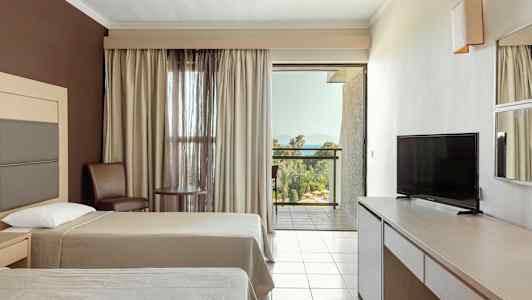 Hotellrom med to senger, TV, bord og stoler samt en balkong med utsikt mot grønne omgivelser og havet. (AI-generert tekst)