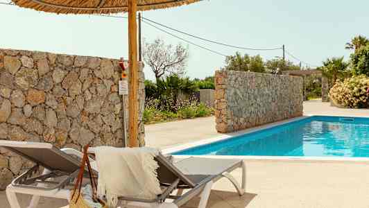 To solsenger med et håndkle og en stråveske ved siden av et basseng. En steinmur og palmer er i bakgrunnen. (AI-generert tekst)