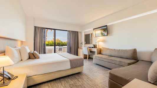 Et hotellrom med en seng, sofaer, skrivebord med speil, og en sittegruppe nær et glassvindu. (AI-generert tekst)