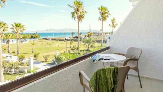 En balkong med bord, stoler og utsikt over en grønn hage med palmer og havet i bakgrunnen. (AI-generert tekst)