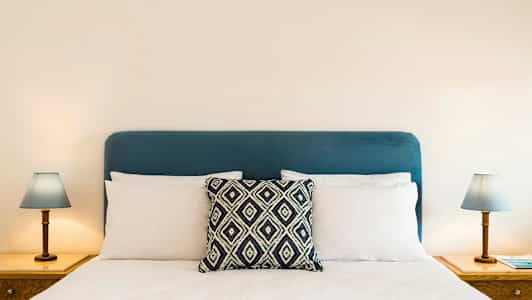 En seng med en blå hodegjerde, hvite puter og en dekorativ pute i midten. To nattbord med lamper på hver side. (AI-generert tekst)