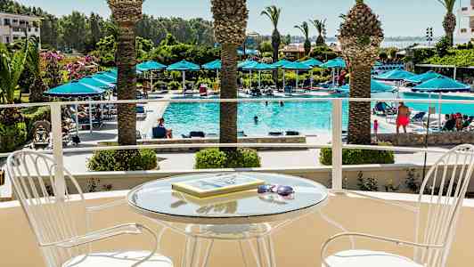 En balkong med et glassbord og stoler, med utsikt over et basseng med solsenger og palmer. (AI-generert tekst)