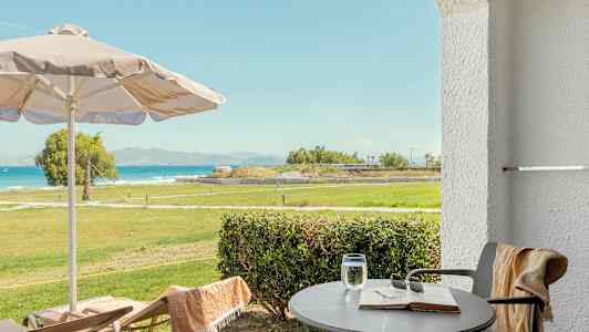 Utsikt fra terrasse med bord, stoler, parasoll og grønt område mot havet. (AI-generert tekst)