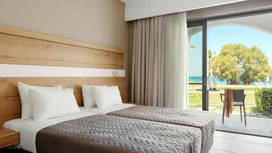 To senger med hvite puter og grå sengeteppe, ved siden av en glassdør som fører til en hage med utsikt over havet. (AI-generert tekst)