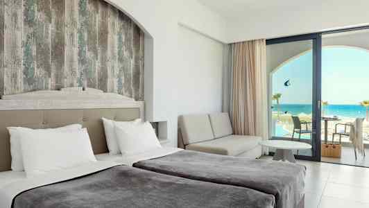 Hotellrom med dobbeltsenger, en sittegruppe og utsikt over havet gjennom en glassdør. (AI-generert tekst)