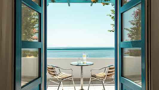 Utsikt gjennom åpne dører mot en terrasse med havutsikt, inkludert et bord og to stoler. (AI-generert tekst)