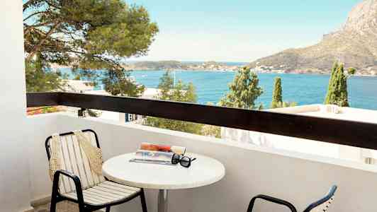 En balkong med stoler og bord med en bok og solbriller på. Utsikt over havet og en fjellaktig bakgrunn. (AI-generert tekst)