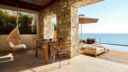 En uteplass med bord, stoler, hengende stol og solsenger ved sjøen. (AI-generert tekst)