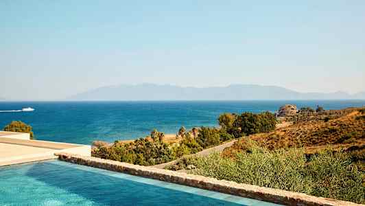 Utsikt over et basseng, kysten og havet med en båt som passerer. Fjellene er synlige i bakgrunnen. (AI-generert tekst)