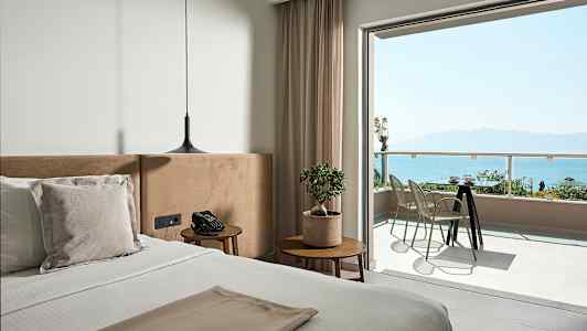 Et rom med en seng, nattbord, moderne hengende lampe og en balkong med utsikt over havet. (AI-generert tekst)