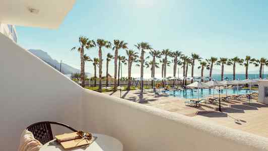 Utsikt fra en balkong mot et basseng med solsenger og palmetrær i solskinn. (AI-generert tekst)