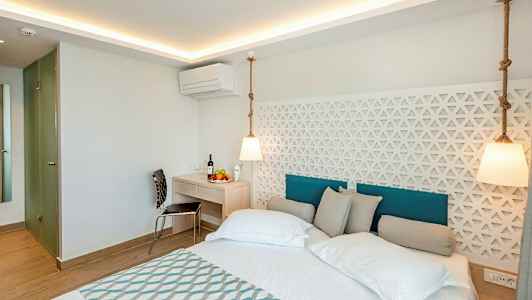 Bildet viser et moderne hotellrom med en dobbeltseng, hvite vegger og en dekorativ panel bak sengen. Et skrivebord med stol og frukt er synlig i rommet. (AI-generert tekst)