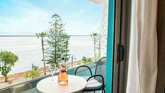 Utsikt fra balkongen med bord, stoler og en flaske rosévin, mot hav og trær. (AI-generert tekst)