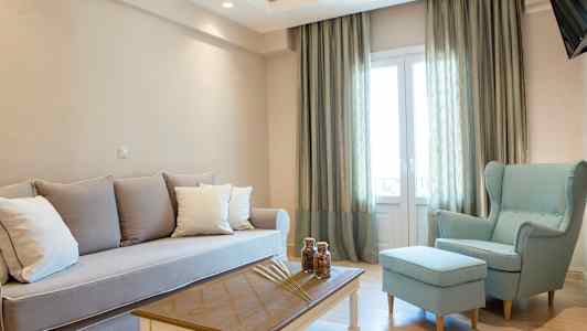 En stue med lyse møbler, inkludert en sofa, en lenestol og et salongbord, ved siden av store vinduer med gardiner. (AI-generert tekst)