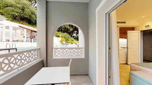 En balkong med et hvitt bord og en stol, utsikt mot bassengområdet og innsikt til et rom. (AI-generert tekst)