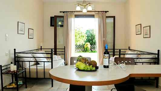 Et innendørs miljø med et bord med frukt og en vinflaske, to senger og en åpen dør som leder til en hage. (AI-generert tekst)