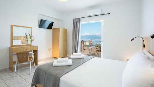 Et lyst hotellrom med en dobbeltseng, skrivebord, speil og en balkong med utsikt over havet. (AI-generert tekst)