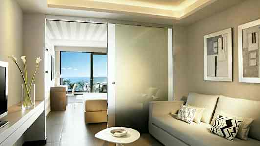 Lyst hotellrom med sofa, lavt bord og en glassvegg mot soveromsdelen med dobbeltseng. Balkong med havutsikt er synlig. (AI-generert tekst)