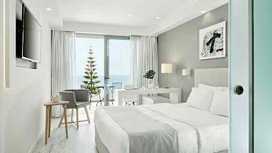 Moderne hotellrom med dobbeltseng, skrivebord og skyvedør ut til en balkong med havutsikt. (AI-generert tekst)