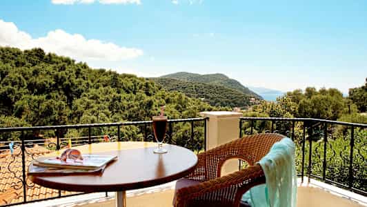 Utsikt fra en balkong med bord, drikke og en stol med et klede samt trær og fjell i bakgrunnen. (AI-generert tekst)