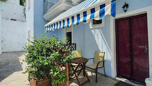 En liten uteplass med et bord, stoler og en markise foran en lyseblå bygning. (AI-generert tekst)