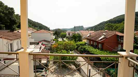 Utsikt fra en balkong over hus med tegltak og grøntområder samt fjell i bakgrunnen. (AI-generert tekst)