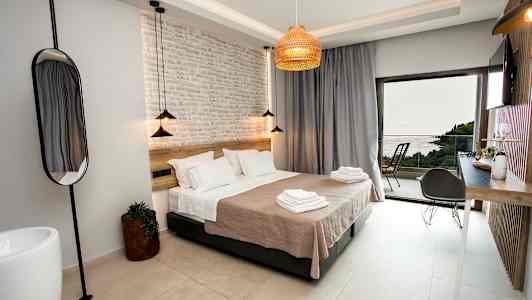 Et moderne hotellrom med en stor seng, lyse vegger og en dør som åpner seg til en balkong med havutsikt. (AI-generert tekst)