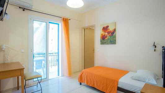 Et enkeltrom med en seng med et oransje teppe, skrivebord og stol, samt en glassdør som fører til en balkong. (AI-generert tekst)