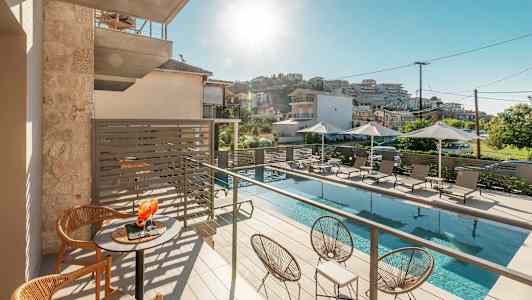 Bilde av en solrik balkong med bord og stoler, et bassengområde nedenfor med solsenger og parasoller. (AI-generert tekst)