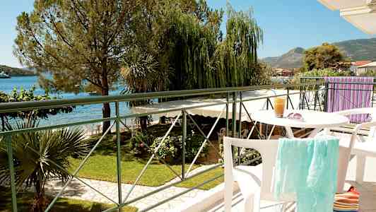 Utsikt fra en balkong med bord og stoler, med grøntområder, sjø og fjell i bakgrunnen. (AI-generert tekst)