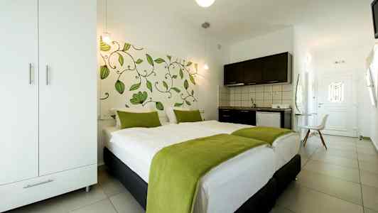 Et hotellrom med en dobbeltseng dekket med hvitt sengetøy og grønne tepper, et klesskap, en kjøkkenkrok og en dør i bakgrunnen. (AI-generert tekst)